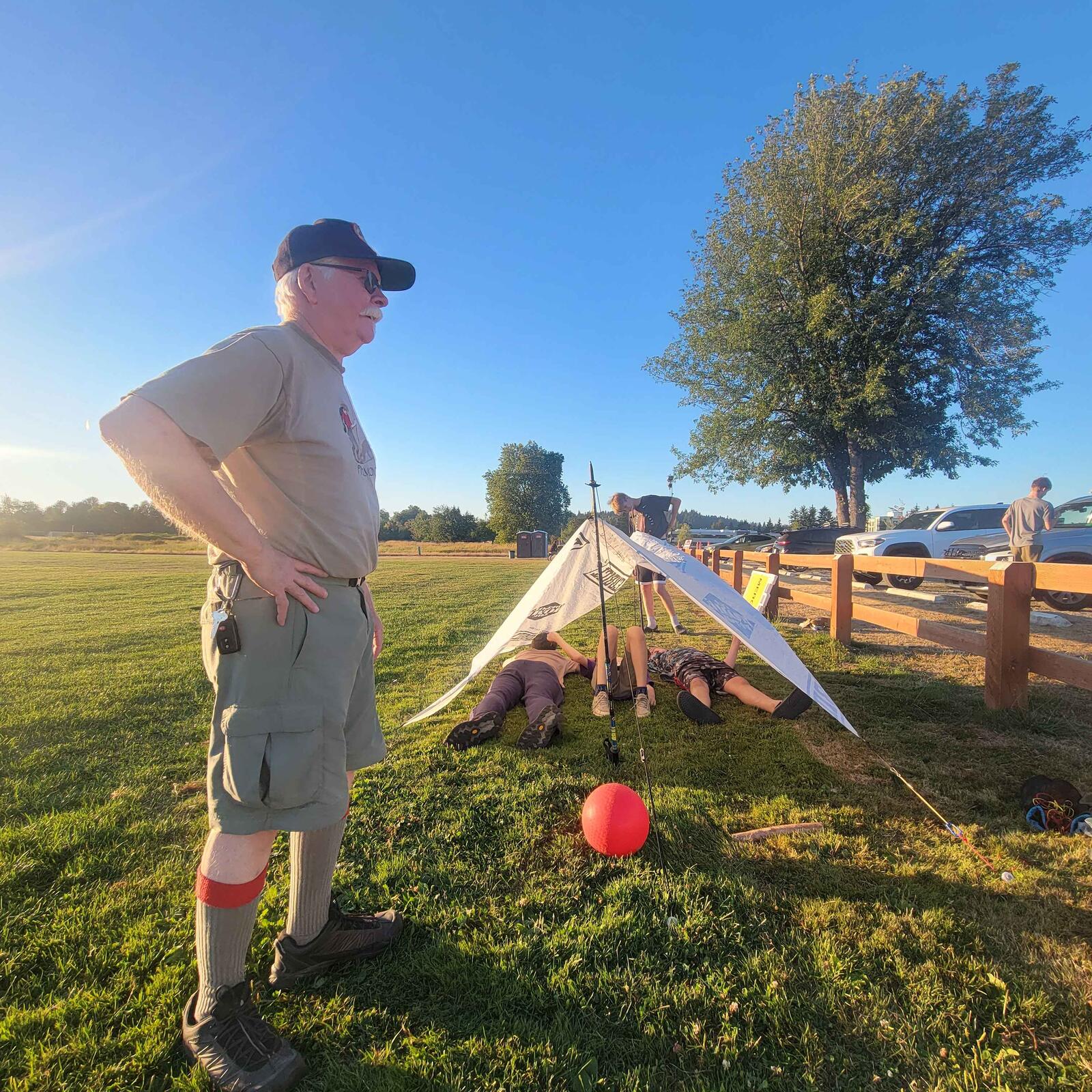 An adult leader watching scouts pitch a tent in the evening light